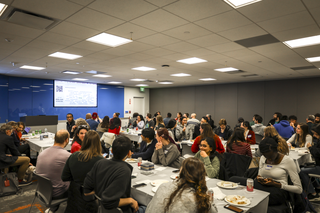 A classroom of students and community members enjoying thanksgiving style food at tables of six or seven.