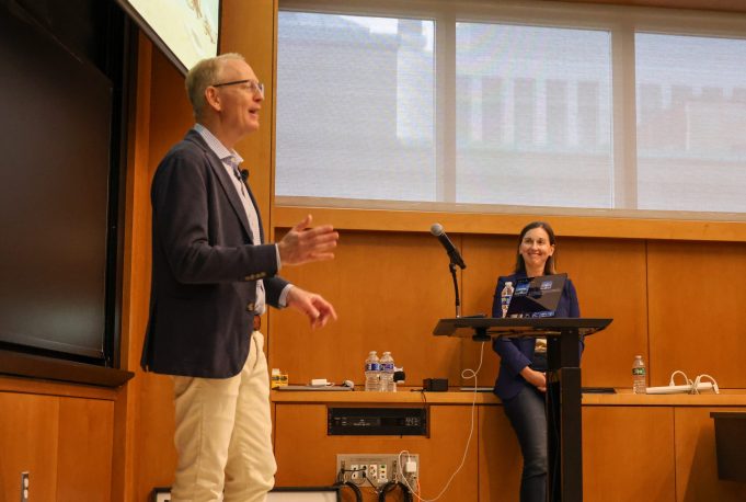 A man speaking at a lecture with a woman standing behind a lectern beside him.