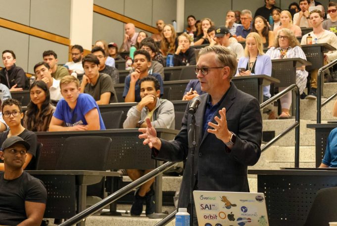 A man speaks to students in a tiered classroom, standing at a microphone.