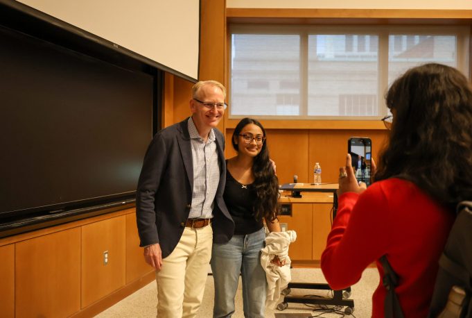 Two people posing for a photo in a classroom as another person takes their picture with a smartphone.