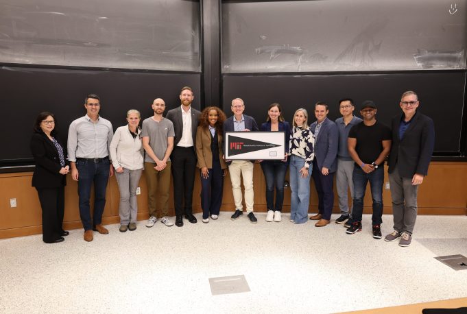 A group of fourteen people stands in a classroom in front of a blackboard, with two people holding a framed pennant from MIT.