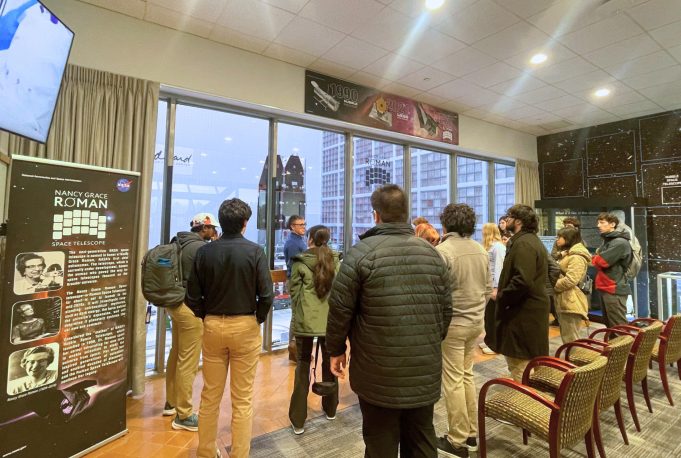 A group of people in a room facing a window with a space-themed exhibit, including a banner about the Nancy Grace Roman Space Telescope. Transcribed Text: Nancy Grace Roman Space Telescope